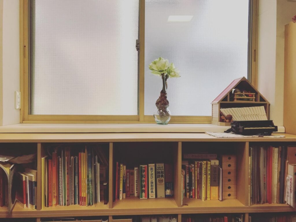 音楽書籍とミニチュアピアノが並ぶ窓辺の本棚。アマリリスの花が飾られている